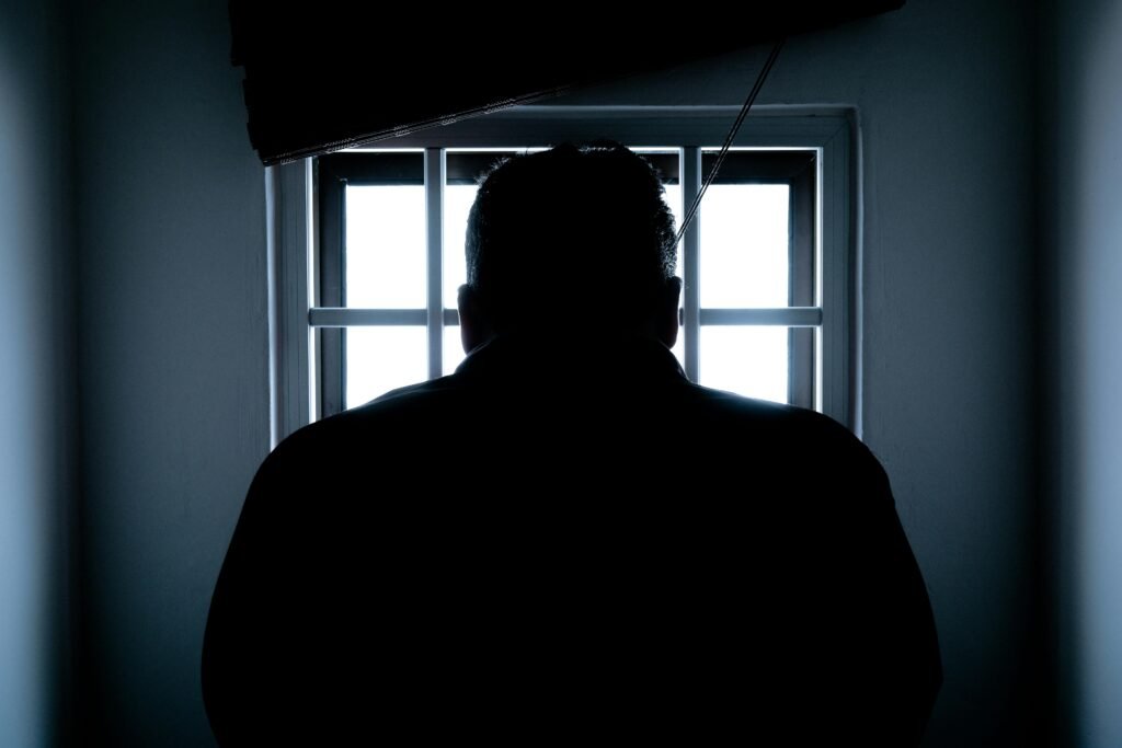 pexels-photo-143580-143580 Dark silhouette of a man in a prison cell, backlit by window bars.
